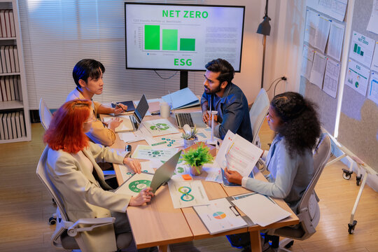 Multiethnic business team discussing ESG sustainability strategy and net zero carbon emission report on screen during meeting with laptops and green documents
