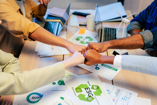 Top view of multiethnic team doing fist bump over ESG sustainability documents and wind turbine model, celebrating teamwork and green business success