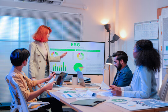 Redhead businesswoman presenting ESG sustainability reporting and rating distribution charts on screen to multiethnic team with laptops and wind turbine model in office