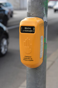 A yellow pedestrian crossing device on a pole with Croatian text meaning Please wait and Pedestrians press for green light