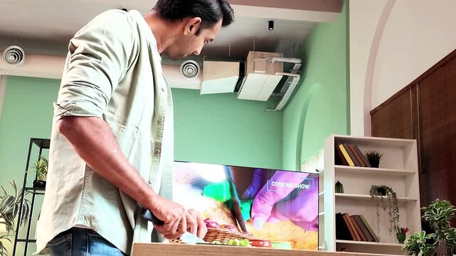 Young Indian man chopping fresh okra in a modern kitchen with a knife while watching a cooking recipe on TV, home cooking, vegetable preparation and everyday lifestyle concept.