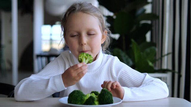 Young girl happily eats broccoli at table, lifting floret from plate and taking bite. Child enjoys fresh green vegetables, offering simple balanced diet and healthy eating example at home.