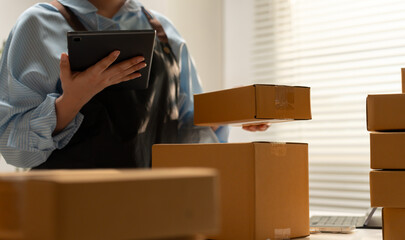 Small business worker holding digital tablet and cardboard parcel among stacked shipping boxes, illustrating ecommerce inventory management, warehouse organization, order logistics operations. © Father_Studio