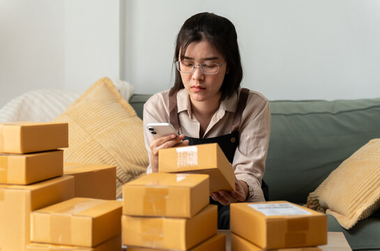 Serious young Asian female store owner holding cardboard parcel box while checking smartphone at desk. Small business seller reviewing shipping address error or inventory stock issue.