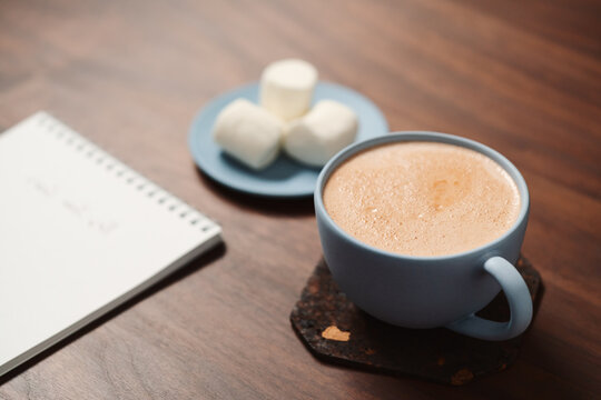 Hot chocolate in a blue cup with marshmallow and notepad on walnut table
