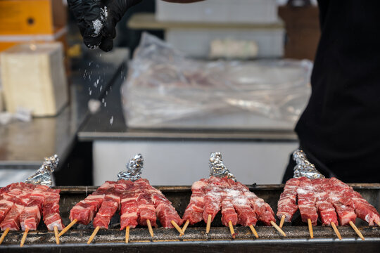 Chef seasons raw meat skewers traditional Italian arrosticini Abruzzesi  with coarse salt over a grill while preparing food in a commercial kitchen or market stall.