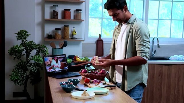 Young Indian man learning to make modak dumplings in a modern kitchen while watching a cooking tutorial on a laptop, homemade food, Indian cuisine and culinary skills concept.