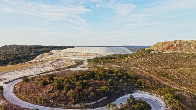 Gravel roads wind through an open mine excavation and scrub-covered hills, with forest along the horizon under a cloudy sky during the day.