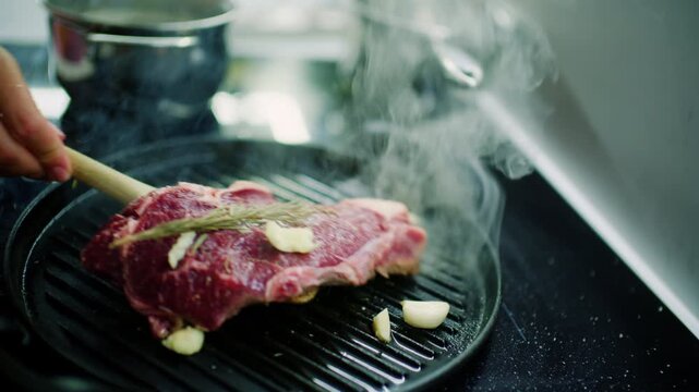 Wooden spatula moving a raw beef steak with rosemary and garlic on a smoking cast iron grill pan, French Riviera, France.