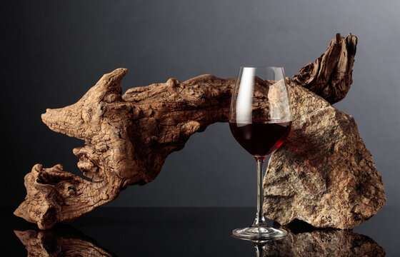 Glass of red wine with old driftwood and a piece of granite rock on a black reflective background.