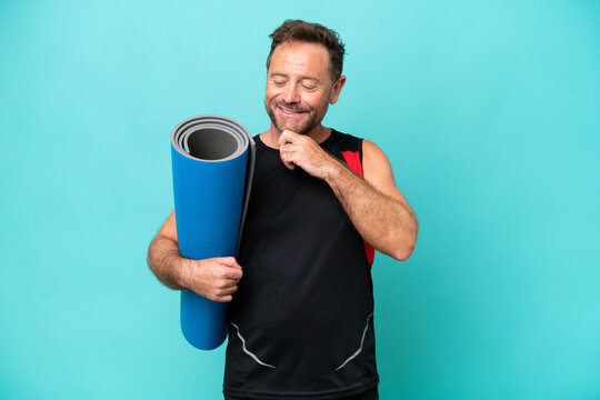 Middle age sport man going to yoga classes while holding a mat isolated on blue background looking to the side and smiling