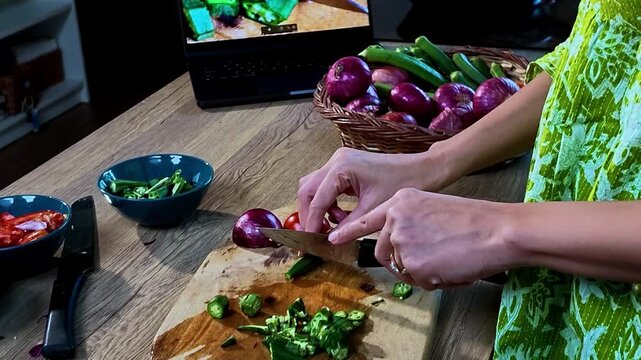 Young Indian woman chopping fresh okra with a knife in a modern kitchen while watching a cooking tutorial on a laptop, home cooking, vegetable preparation and everyday lifestyle concept.