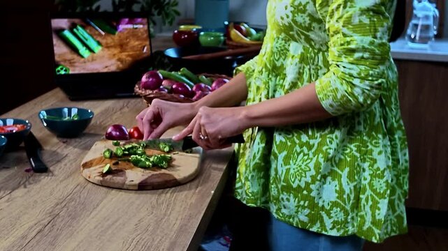 Young Indian woman chopping fresh okra with a knife in a modern kitchen while watching a cooking tutorial on a laptop, home cooking, vegetable preparation and everyday lifestyle concept.