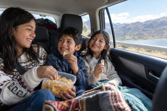Three happy hispanic siblings sharing chips in back seat of car on road trip with mountains, lake view, family travel