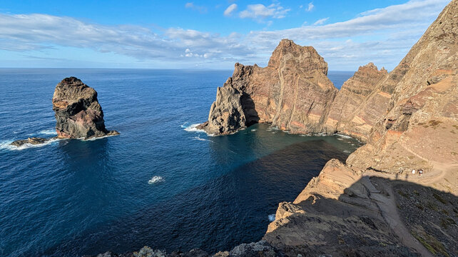 Layered red and ochre volcanic cliffs and a sea stack rising from deep blue Atlantic water, with hikers on the coastal trail, at Ponta do Castelo, Ponta de S&atilde;o Louren&ccedil;o, Madeira, Portugal.