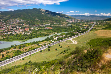High angle panoramic view of asphalt highway road with traffic along green Kura River valley and Caucasus mountains near Mtskheta Georgia under a bright blue sky © Vladimir Bikhovskiy