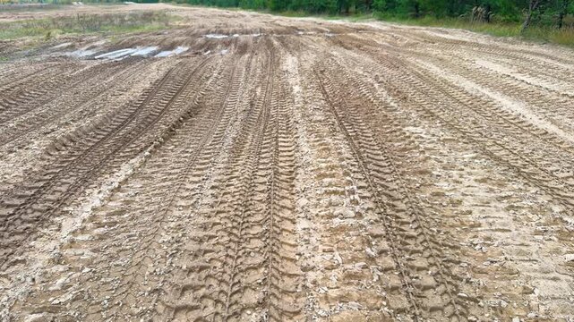 Parallel tire tracks cross a muddy gravel field with shallow puddles beside a forest, wet ground texture, vehicle imprints, rural terrain
