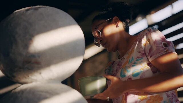Focused latina artisan meticulously crafting a large paper mache snowman sculpture in her workshop, applying paste with a brush in the sunlight