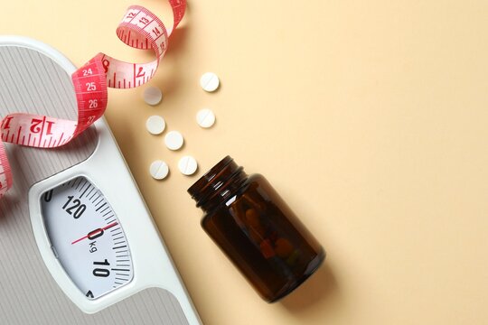 Scales with weight loss pills and measuring tape on beige background, top view