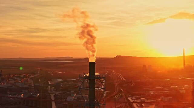 Smoke rises from a factory smokestack above buildings and roads in an industrial district as the sun sets over the horizon.