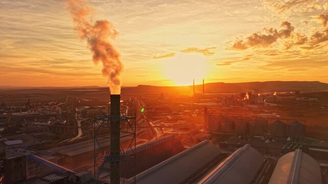 Factory buildings and tall chimneys release smoke across an industrial area as the sun sets on the horizon, with city buildings visible in the distance.