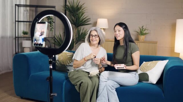 Young asian makeup artist and elderly caucasian woman creating online content, reviewing cosmetic products, and broadcasting a live stream
