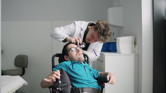 Eye doctor examining a patient with spina bifida sitting in an electric wheelchair. An ophthalmologist performing an eye exam in a hospital room