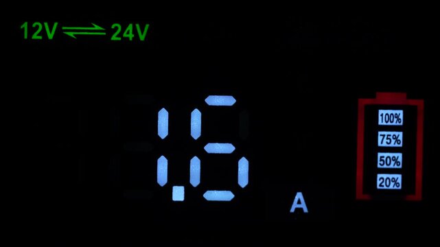 Close up of smart battery charger electronic screen with blue voltage digits 14.6 V and green 12V 24V indicator. Modern diagnostic equipment display in darkness