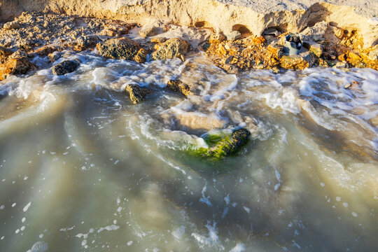 Close-up view of foamy waves washing over algae-covered rocks along sunlit, eroded shoreline. Aruba.