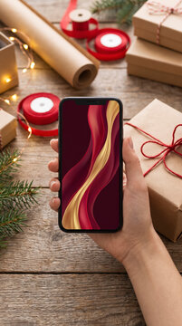 Meticulous gift wrapper holding a smartphone amid kraft parcels, red ribbon spools and fairy lights