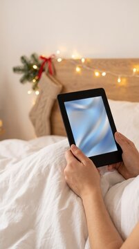 Bedtime reader holding an e-reader under a white duvet with a hanging stocking and fairy lights