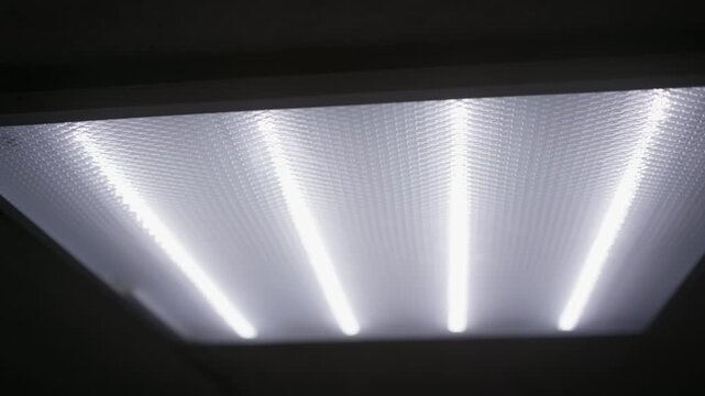 Fluorescent ceiling light flickers in dark interior room. Low angle view captures the textured diffuser panel illuminated by bright tube bulbs in a gloomy building hallway.