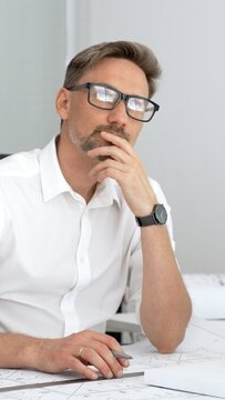 Male architect planning new building project in office, looking up and thinking, developing creative ideas, working on blueprints with laptop