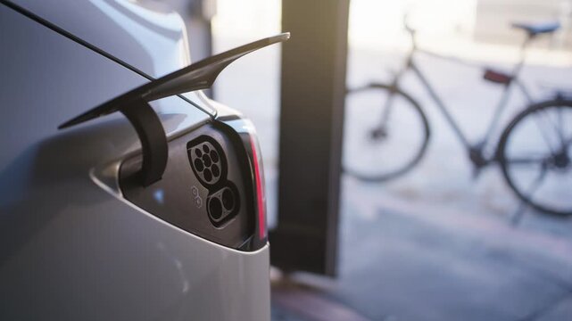 Person unplugs charging cable from electric car in home garage as port lid automatically closes. Soft sunlight illuminates the driveway while a bicycle sits softly blurred in the background.