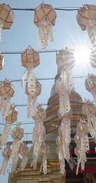 White hanging lanterns with ornate streamers frame golden stupa and sunburst at Wat Phra That Hariphunchai, Lamphun, Thailand, during 2026 lantern festival, vertical view beneath blue sky