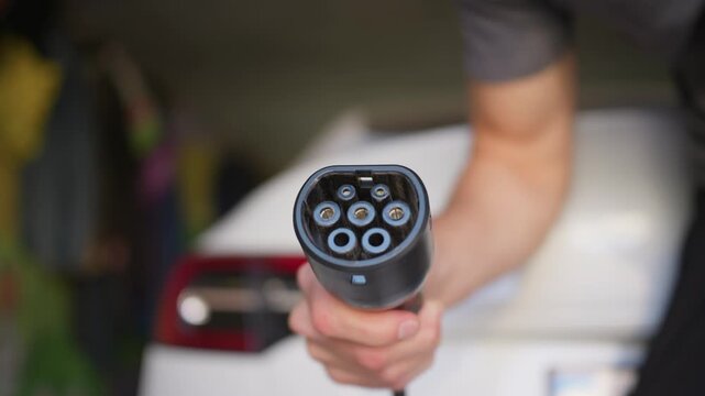 Male hand holds electric vehicle charging plug in home garage. Selective focus emphasizes the black power connector pins against a blurred automobile in soft daylight.