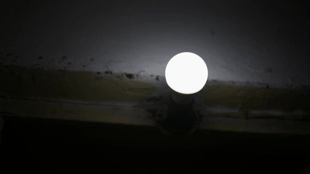 Glowing light bulb shines in dark interior room on ceiling against concrete. Direct illumination creates intense high contrast against deep surrounding shadows.