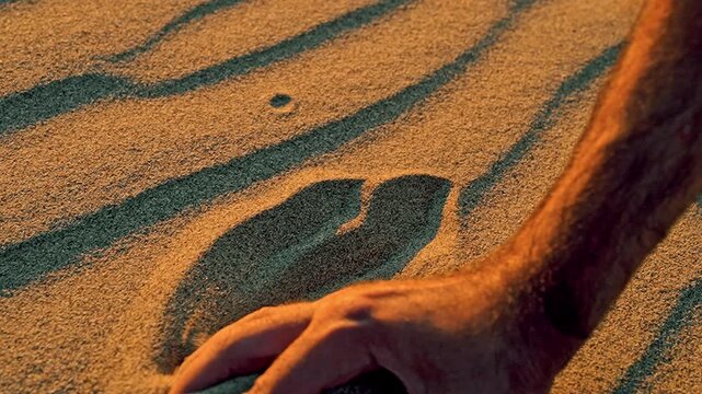 Slow motion footage of a man scooping up a handful of sand and releasing it from his palm as the wind carries the grains through the air.