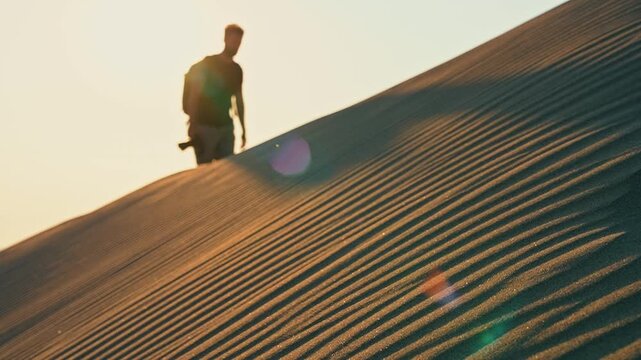 Desert sand dunes reveal a traveler walking in slow motion as wind lifts drifting sand grains and sunlight illuminates the landscape.
