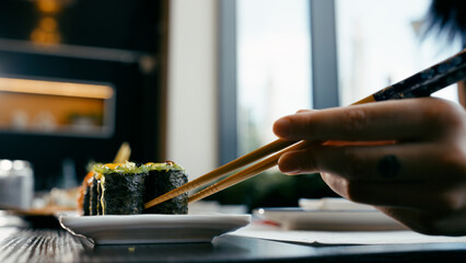 Hand using chopsticks to pick up a maki sushi roll from a plate in a Japanese restaurant setting. Fresh seafood meal © DMegias