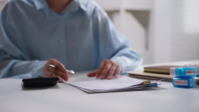 Office worker holding a pen while checking printed financial reports beside a calculator, illustrating bookkeeping, expense verification, accounting accuracy, and routine business administration.