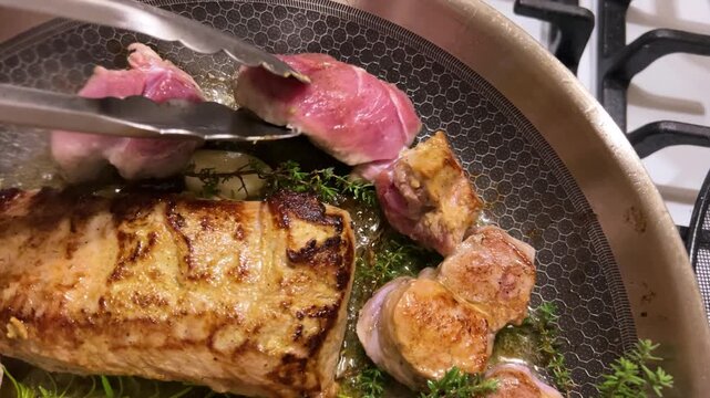 Chef cooks tender veal medallions with garlic and aromatic thyme branches on a frying pan as fresh herb-infused oil sizzles around gourmet steaks in a restaurant kitchen close-up.