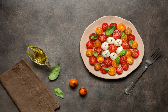 Caprese salad with mozzarella, colored cherry tomatoes, basil leaves, and olive oil on a dark grunge background; top-down view, flat lay.