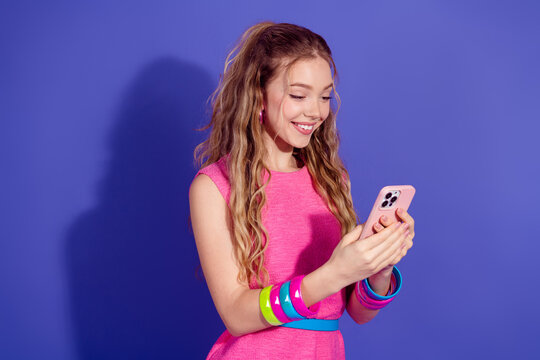 Young woman wearing a trendy pink outfit using a smartphone, standing against a purple background, smiling with joy