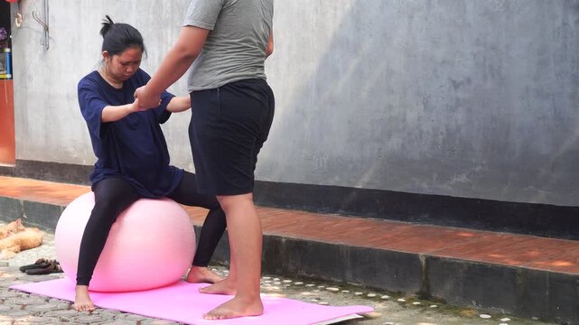 Asian woman exercising on a pink stability ball outdoors supported by partner.