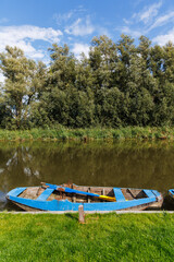 Weathered blue wooden rowing boat with oars moored on a calm canal beside green grass, tall willows reflected in the water under a blue sky. © Ostrovskajah