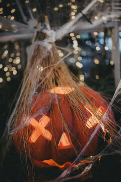 Close shot of illuminated carved Halloween pumpkin with witch broom bristles and cobwebs against bokeh light.