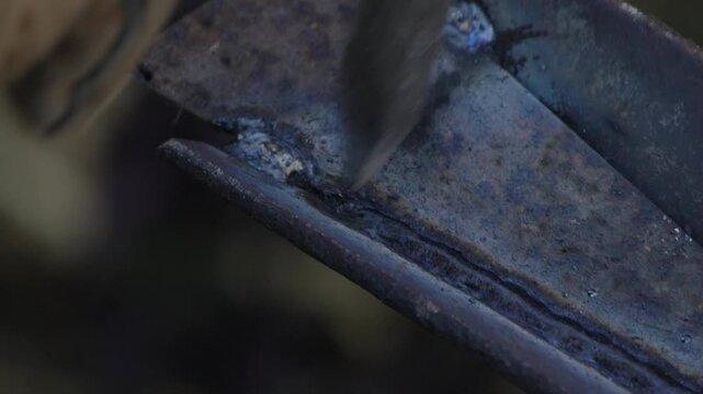Metal slag removal occurs as a worker chips away excess material from a weld joint on steel. Close-up macro shot.