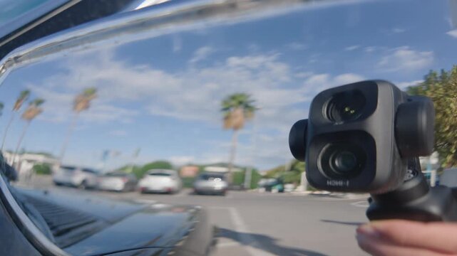 Close-up reflection of a hand holding a compact gimbal camera in a chrome car mirror, with palm trees and blue sky, French Riviera, France.