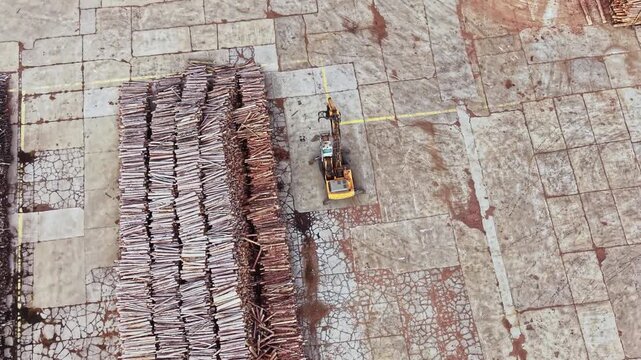 An excavator works beside long rows of cut timber in a paved yard, viewed from above during the day.